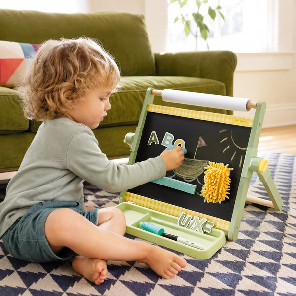 A toddler sits on a patterned rug in a living room, using a wooden children's magnetic writing board and tray. The child is writing letters on the blackboard surface, which also has a yellow textured brush and a white roll at the top.