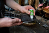 Person applying a green bottle of shoe polish to a black shoe in a workshop setting.