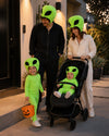 A family of four dressed in matching neon green alien costumes, standing outside a house. The father wears a black costume, the mother wears a cream-colored costume, the young girl holds an orange pumpkin bucket, and the baby is seated in a black stroller.
