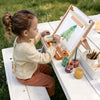 A young child sitting outdoors at a white wooden table, painting on a small easel with colorful paint jars and brushes nearby.