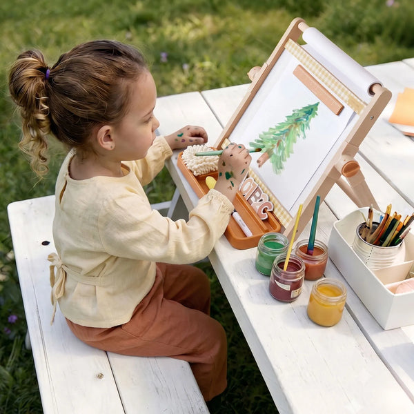 A young child sitting outdoors at a white wooden table, painting on a small easel with colorful paint jars and brushes nearby.