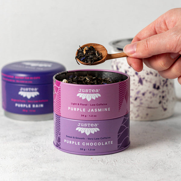 Hand holding a wooden spoon with tea leaves over a Justea purple tea canister.