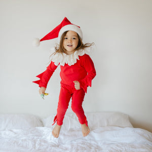 Child wearing a red elf pajama costume sitting on a white bed during the holidays