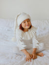 Child wearing a white outfit with ruffled details sitting on a white bed.