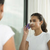 Woman applying facial mask with a purple brush in front of a mirror