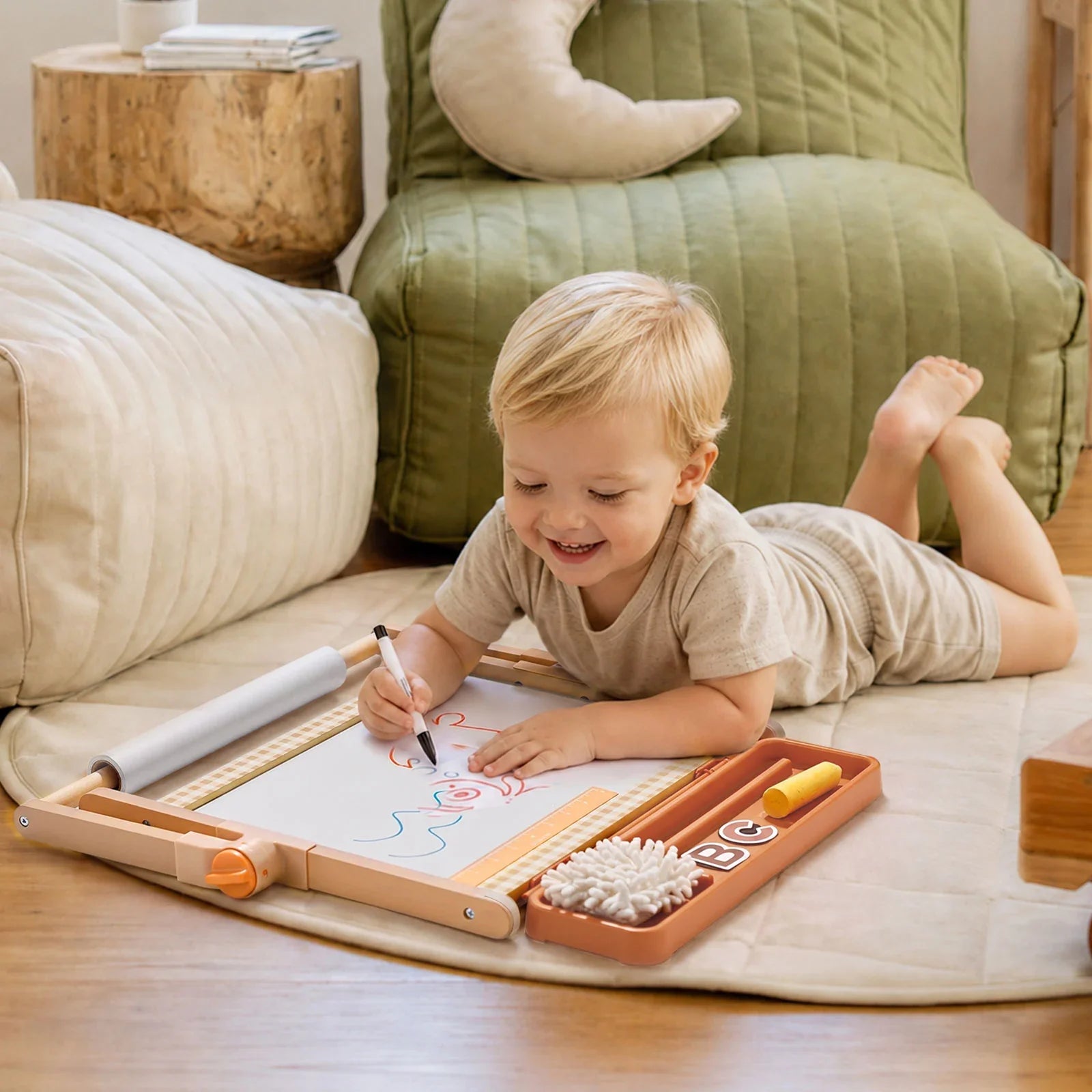 A toddler lies on a cream rug in a cozy living room, drawing on a wooden magnetic activity board with a tray of colorful crayons and cotton swabs.