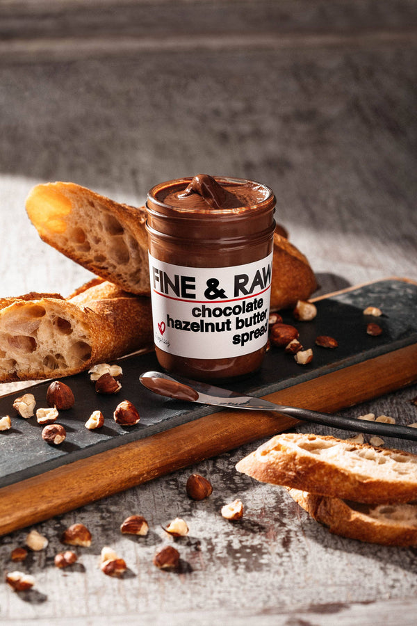 Jar of Fine & Raw chocolate hazelnut butter spread with bread and nuts on a wooden board.