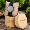 Watch with blue strap on a textured pillow, surrounded by greenery and wooden box with engraved text.