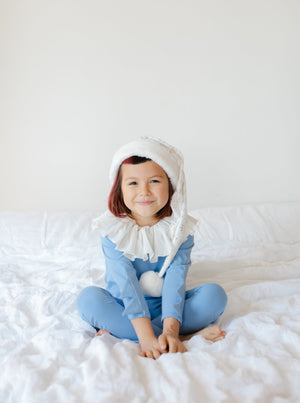 Child wearing a blue Hanukkah elf pajama costume with a white elf hat, sitting on a white bed.