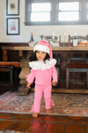 Child in a pink outfit with a Santa hat standing in a room with wooden furniture and a window.