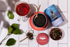 Tea-making setup with a mug of red tea, loose tea leaves, and Justea containers on a white surface.