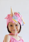 Child wearing a pink unicorn hat with colorful tassels and a gold horn on a light gray background