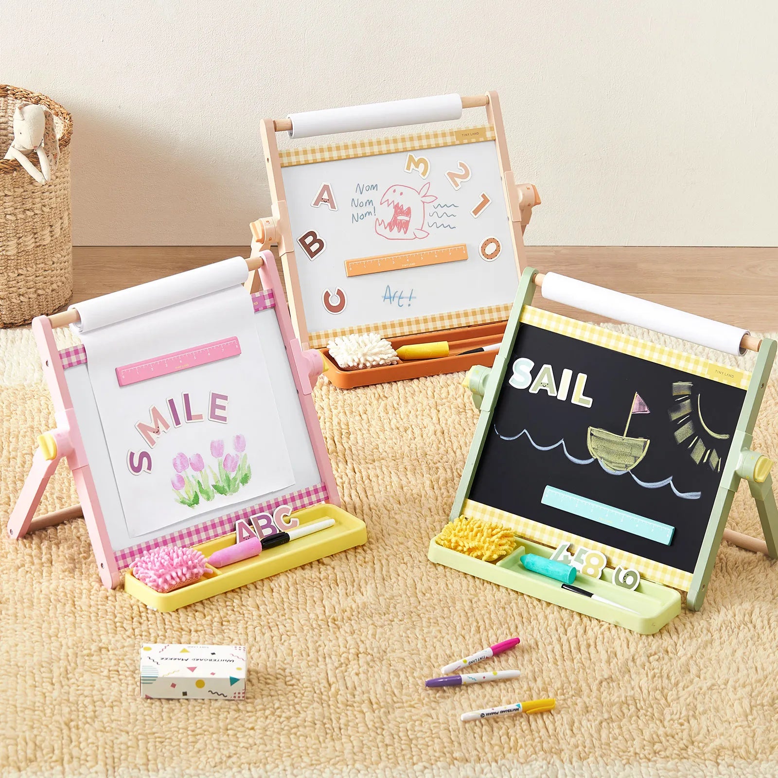 Three small children's magnetic drawing easels arranged on a beige rug. One easel is pink and white with a 'SMILE' board and tulips, one is light wood with alphabet letters, and one is black with a sailboat drawing.