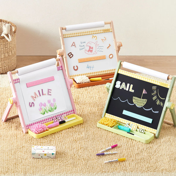 Three small children's magnetic drawing easels arranged on a beige rug. One easel is pink and white with a 'SMILE' board and tulips, one is light wood with alphabet letters, and one is black with a sailboat drawing.