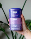 Hand holding three purple tins of Justea tea with plant in background