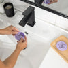 Person holding a purple soap dish with a floral design over a sink, with another soap dish on a wooden tray in the foreground.
