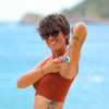Woman applying sunscreen on a beach with a clear blue sky and ocean in the background. NANAH Natural Mango deodorant — aluminum-free, 24-hour protection for normal skin. Mango butter, magnesium & zinc ricinoleate. Plastic-free & family safe. Shop now.