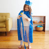 Child in a blue royal costume with a crown and wand, standing in a room with a yellow chair and wooden shelf.