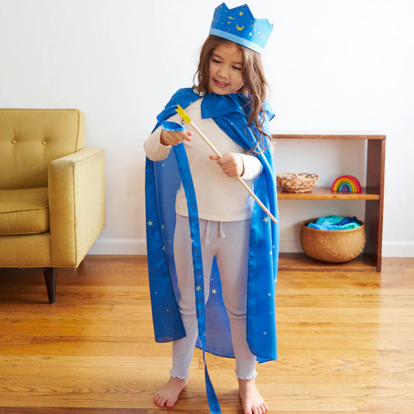 Child in a blue royal costume with a crown and wand, standing in a room with a yellow chair and wooden shelf.