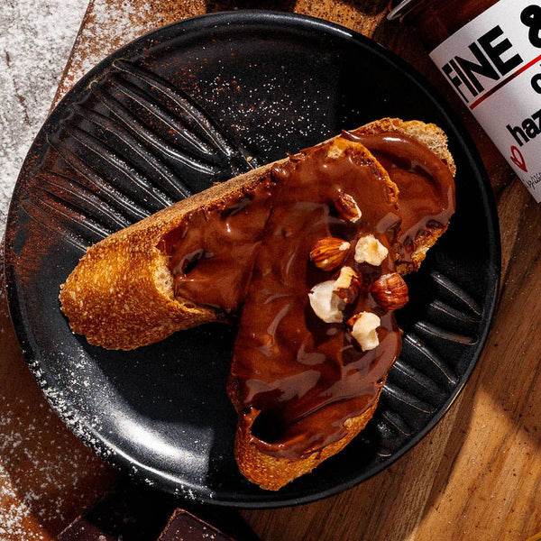 Two slices of bread with chocolate spread and nuts on a black plate, with a bottle of chocolate syrup in the background.