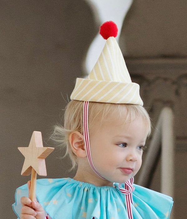Child wearing a clown hat with a pom-pom and holding a star wand, against a neutral background.