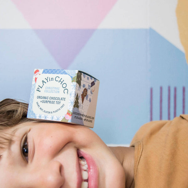 Child with a PLAYin CHOC organic chocolate surprise toy packaging on their face against a colorful geometric background