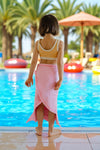 Child in a yellow top and pink skirt standing by a pool with palm trees in the background