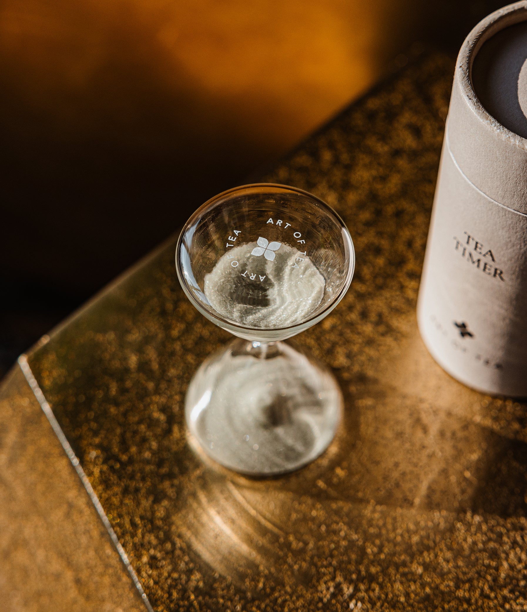 A clear glass tea timer hourglass with printed markings sits on a reflective gold-toned surface next to a white cylindrical tea tin labeled 'TEA TIMER'.