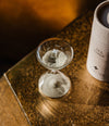 A clear glass tea timer hourglass with printed markings sits on a reflective gold-toned surface next to a white cylindrical tea tin labeled 'TEA TIMER'.