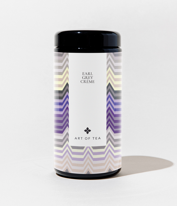 A cylindrical canister of Art of Tea Earl Grey Crème tea, featuring a black lid and a white label with purple, gray, and beige chevron patterns on the sides.