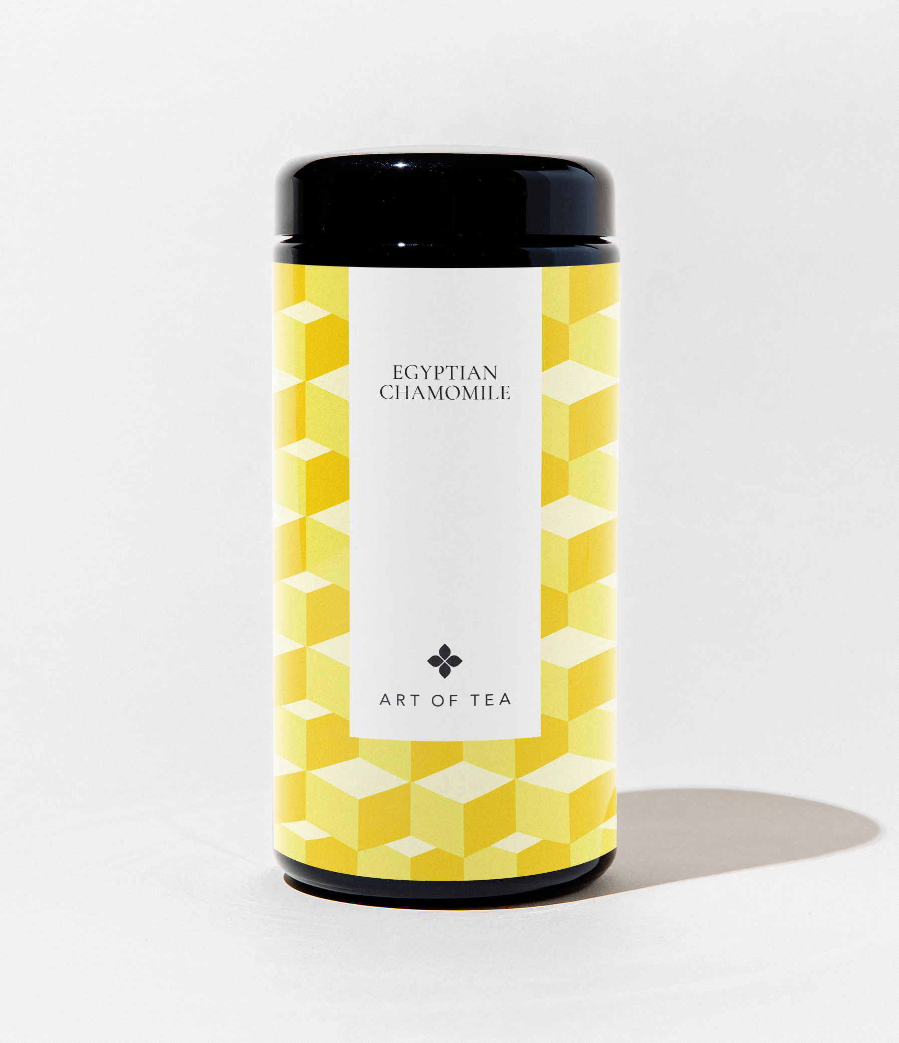 A cylindrical canister of Art of Tea Egyptian Chamomile tea, featuring a black lid and base with a yellow and white geometric cube pattern on the label, set against a plain white background.
