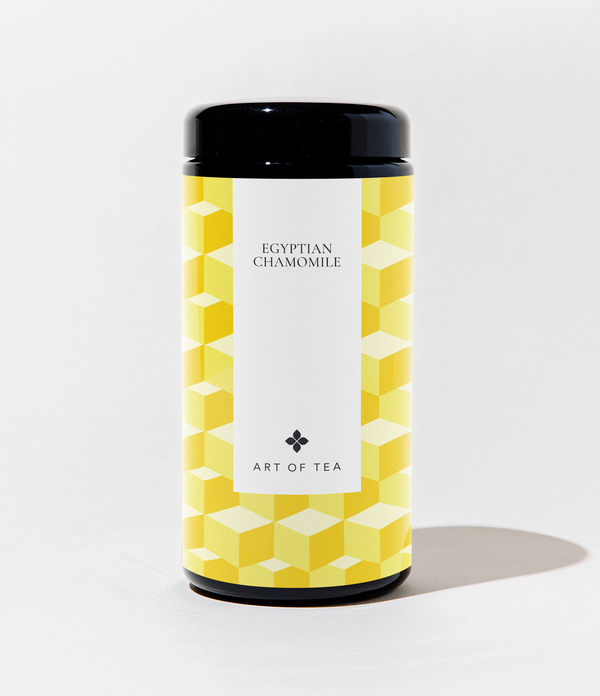 A cylindrical canister of Art of Tea Egyptian Chamomile tea, featuring a black lid and base with a yellow and white geometric cube pattern on the label, set against a plain white background.