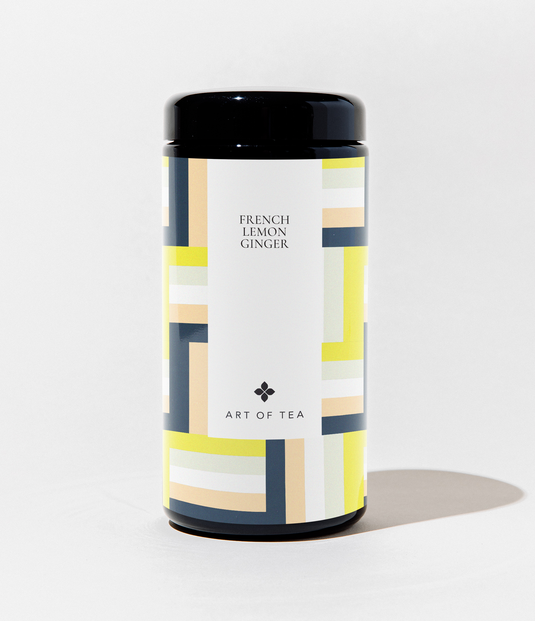 A cylindrical black canister of Art of Tea French Lemon Ginger tea, featuring a white label with a geometric pattern of yellow, navy, and beige blocks, set against a clean white background.