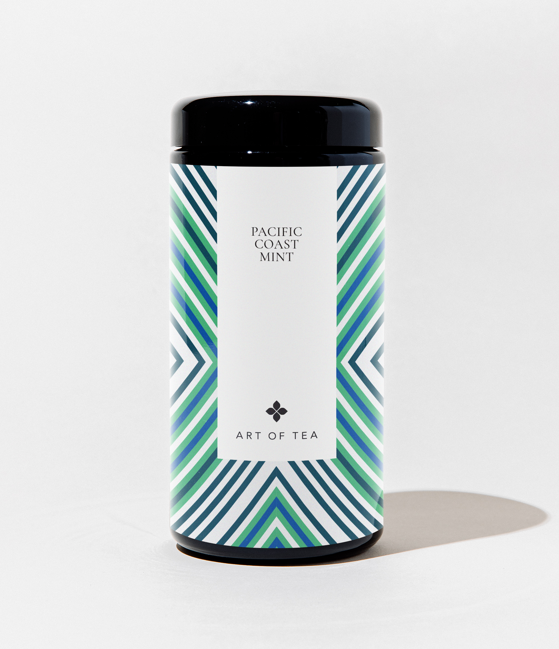 A cylindrical canister of Art of Tea Pacific Coast Mint tea, featuring a black lid and base with a white label and a blue and green geometric pattern on the sides, set against a plain white background.