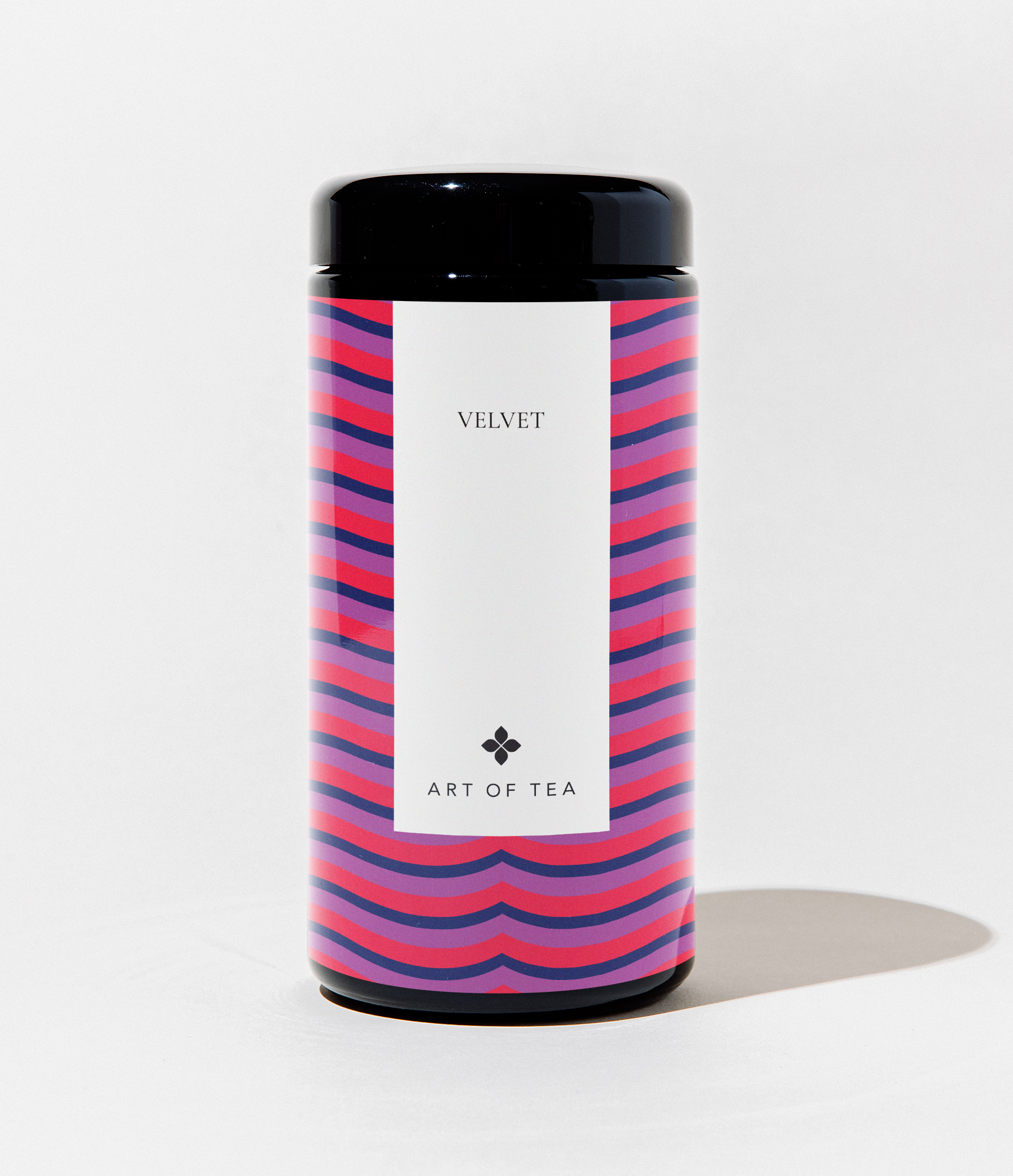 A cylindrical canister of Art of Tea Velvet tea, featuring a black lid and base, and a white label in the center. The canister is decorated with a vibrant pink, purple, and blue wavy stripe pattern on the sides, set against a clean white background.