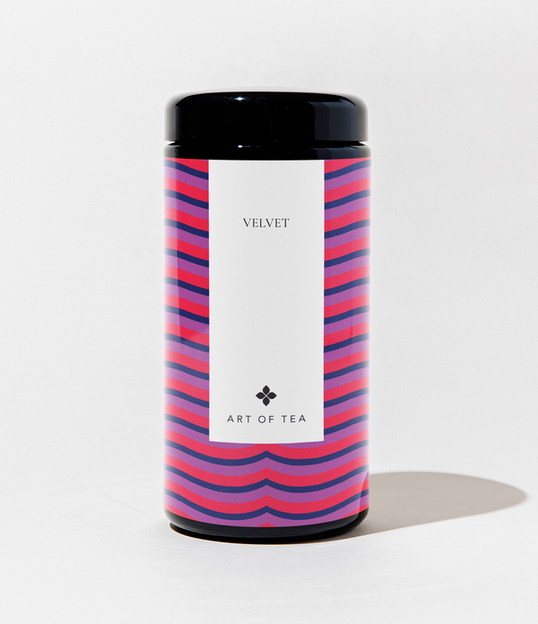 A cylindrical canister of Art of Tea Velvet tea, featuring a black lid and base, and a white label in the center. The canister is decorated with a vibrant pink, purple, and blue wavy stripe pattern on the sides, set against a clean white background.