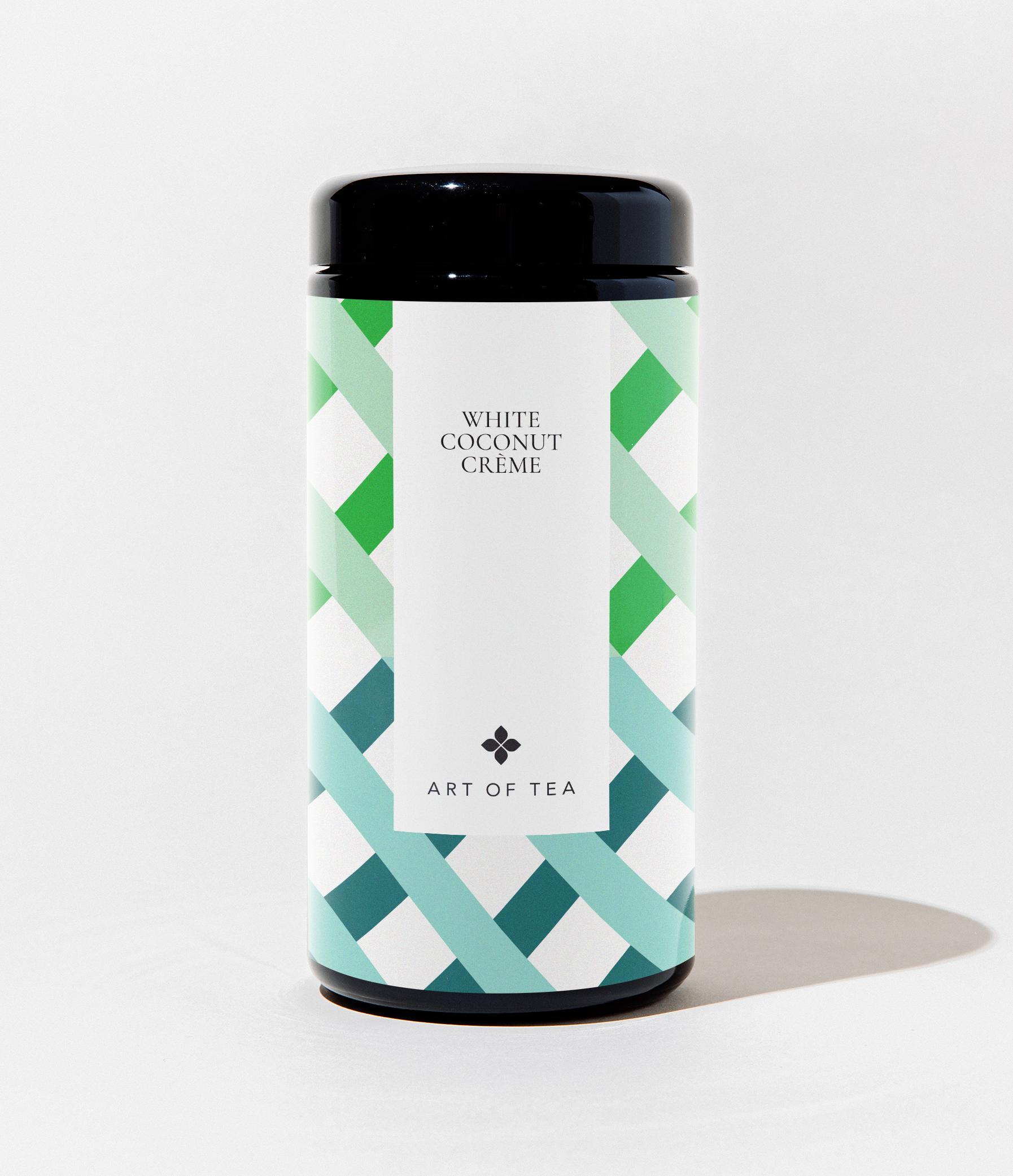 A cylindrical canister of Art of Tea White Coconut Crème tea, featuring a black lid and base with a white label decorated in a green and teal geometric pattern.