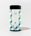 A cylindrical canister of Art of Tea White Coconut Crème tea, featuring a black lid and base with a white label decorated in a green and teal geometric pattern.