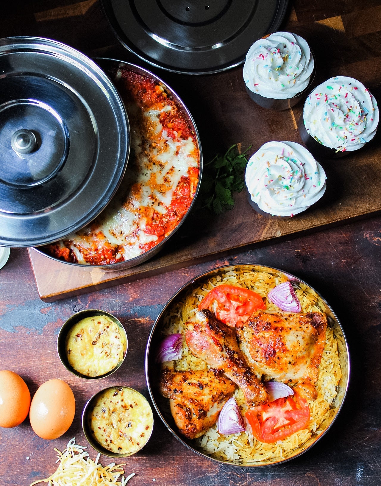 Dishes of food including a pizza, cupcakes, and a dish with chicken and tomatoes on a wooden table.