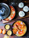 Dishes of food including a pizza, cupcakes, and a dish with chicken and tomatoes on a wooden table.