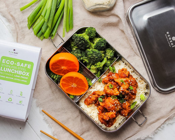 Lunchbox with food including chicken, rice, broccoli, and oranges, next to an Eco Safe container and packaging.