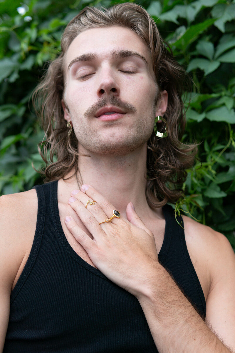 Man with long hair and a beard, wearing a black tank top, against a green leafy background.