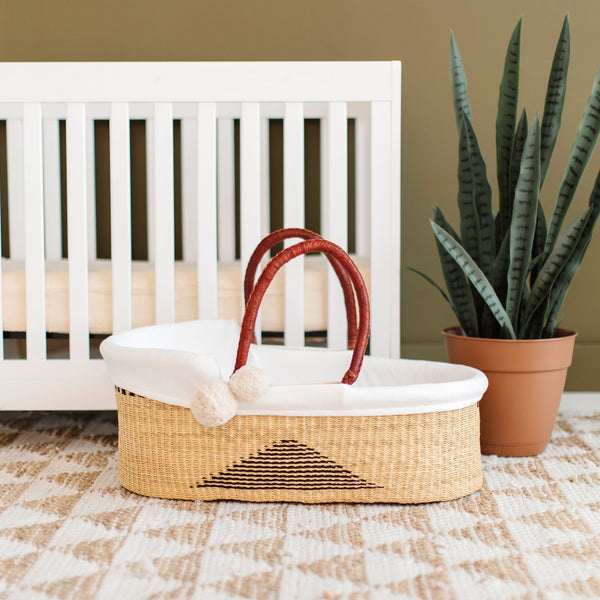 Wicker Moses basket with white cushion in front of a white crib with a potted plant on a checkered floor.