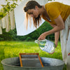 Woman pouring liquid from a bottle into a metal tub outdoors with clothes hanging in the background