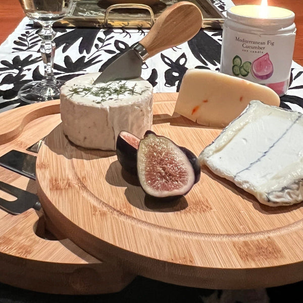 Wooden cheese board with assorted cheeses and figs on a table with a candle in the background.