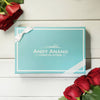 Blue chocolate box with 'Andy Anand Chocolatier' branding, surrounded by red roses on a wooden surface.