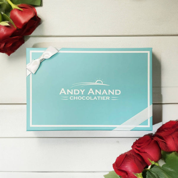 Blue chocolate box with 'Andy Anand Chocolatier' branding, surrounded by red roses on a wooden surface.