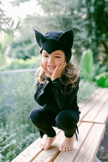Child wearing a black cat costume outdoors on a wooden bench.