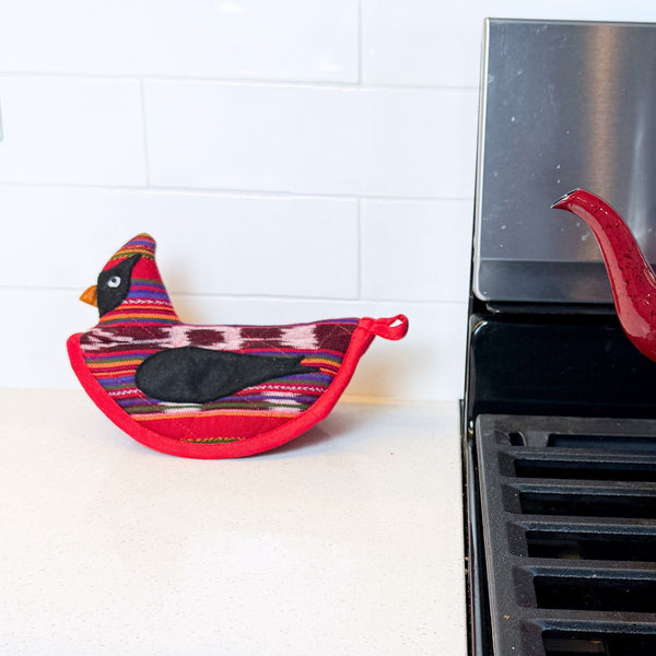 Colorful bird-shaped pot holder on a stove against a white tiled wall.