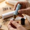 Feminine hands applying amber glass roller bottle of Apotheke DuJardin Cuticle and Nail Oil with light blue label to manicured nails